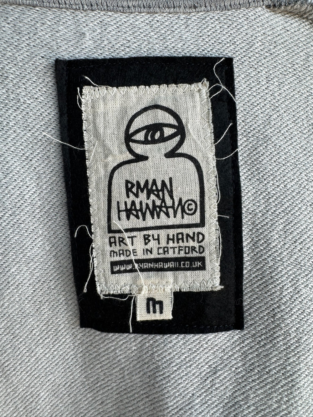 Detailed close-up of the Rman Hawaii garment label featuring a hand-drawn eye graphic, text reading ‘Art by Hand, Made in Catford,’ and website link www.rmanhawaii.co.uk, highlighting artisanal craftsmanship and branding