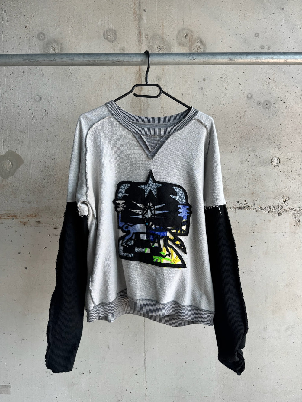 Distressed grey sweatshirt with black contrasting sleeves and bold abstract skull patch featuring layered fabric details in black, blue, and yellow, creating a striking streetwear aesthetic