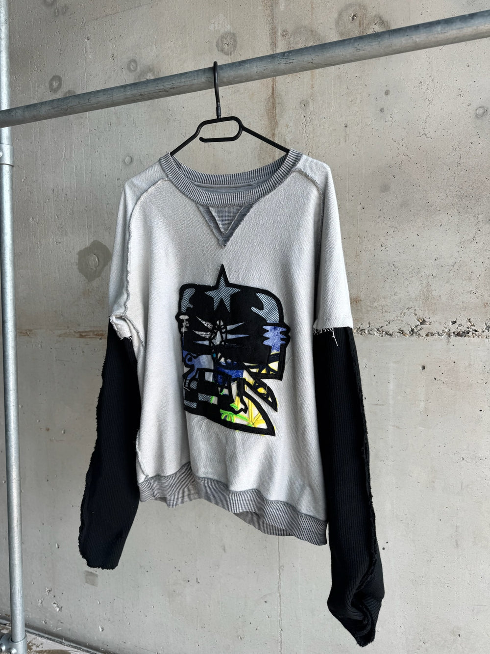 Side angle view of grey distressed sweatshirt with black ribbed sleeves and bold abstract skull patch featuring layered fabric details in blue, yellow, and black for an edgy streetwear look