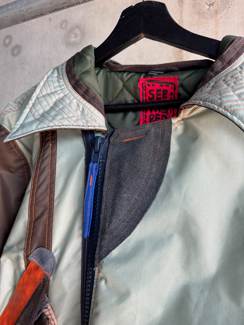 Detail view of a mint green and dark brown parka coat with quilted collar stitching, bright blue zipper, and contrasting red label reading “I SEE RED”; photographed against a raw concrete wall background on a black hanger.