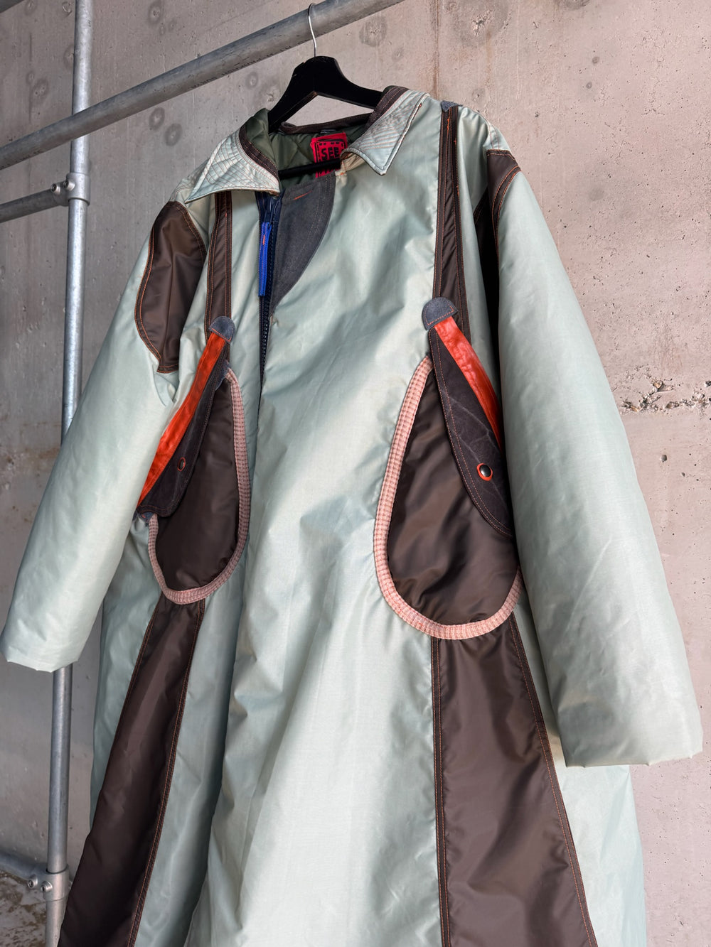 Close-up of a mint green and dark brown unisex parka coat with oversized sculptural flap pockets, orange and grey detailing, and a contrasting zipper; industrial background with concrete wall and metal clothing rack.