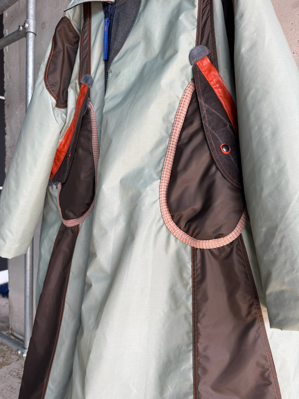 Close-up of a mint green and dark brown oversized parka featuring large teardrop-shaped pockets with red leather accents, pink ribbed edging, and a blue zipper; displayed on a hanger against a raw concrete wall.