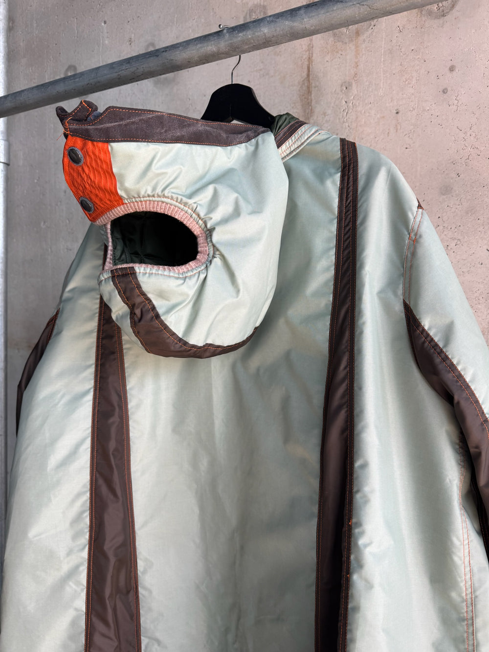 Close-up of the hood on a mint green and brown oversized parka, highlighting the structured design with a quilted orange patch, black snap buttons, and ribbed trim, against an industrial concrete background.