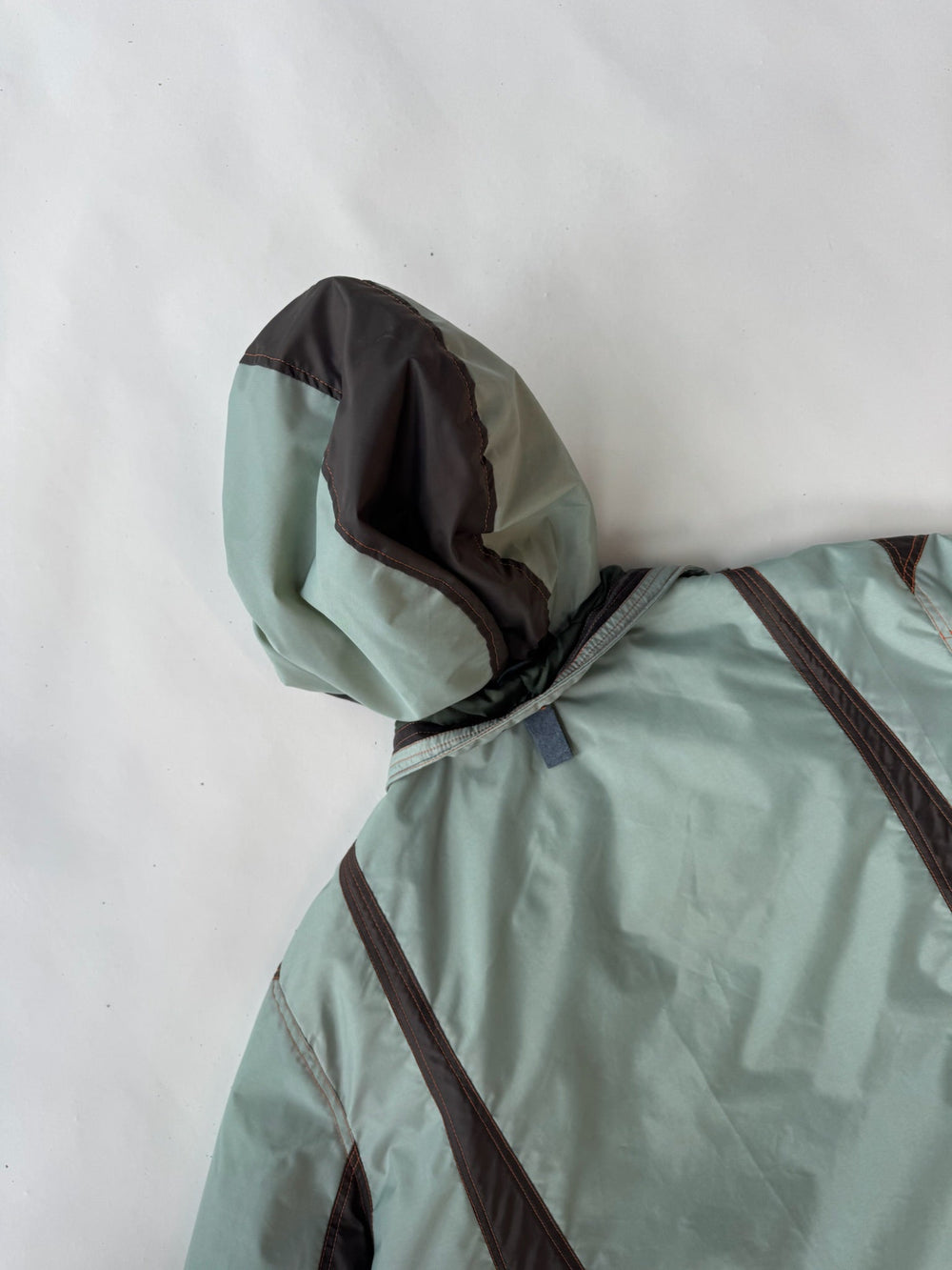 Detail of a sleeve on a mint green and brown technical coat, showcasing the contrast paneling, topstitching, and durable fabric construction, laid flat on a white background.