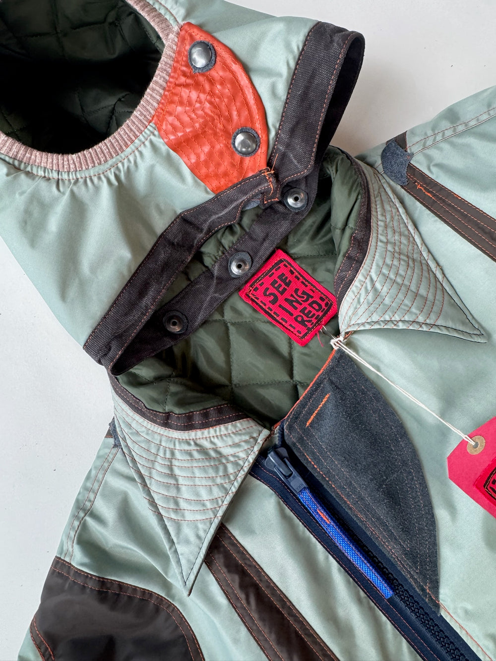 Close-up of a mint green hooded parka with brown and red color-block detailing, exposed blue zipper, quilted stitching on the collar, and a bold red “I SEE IN RED” tag hanging from the neckline, laid flat on a white surface.