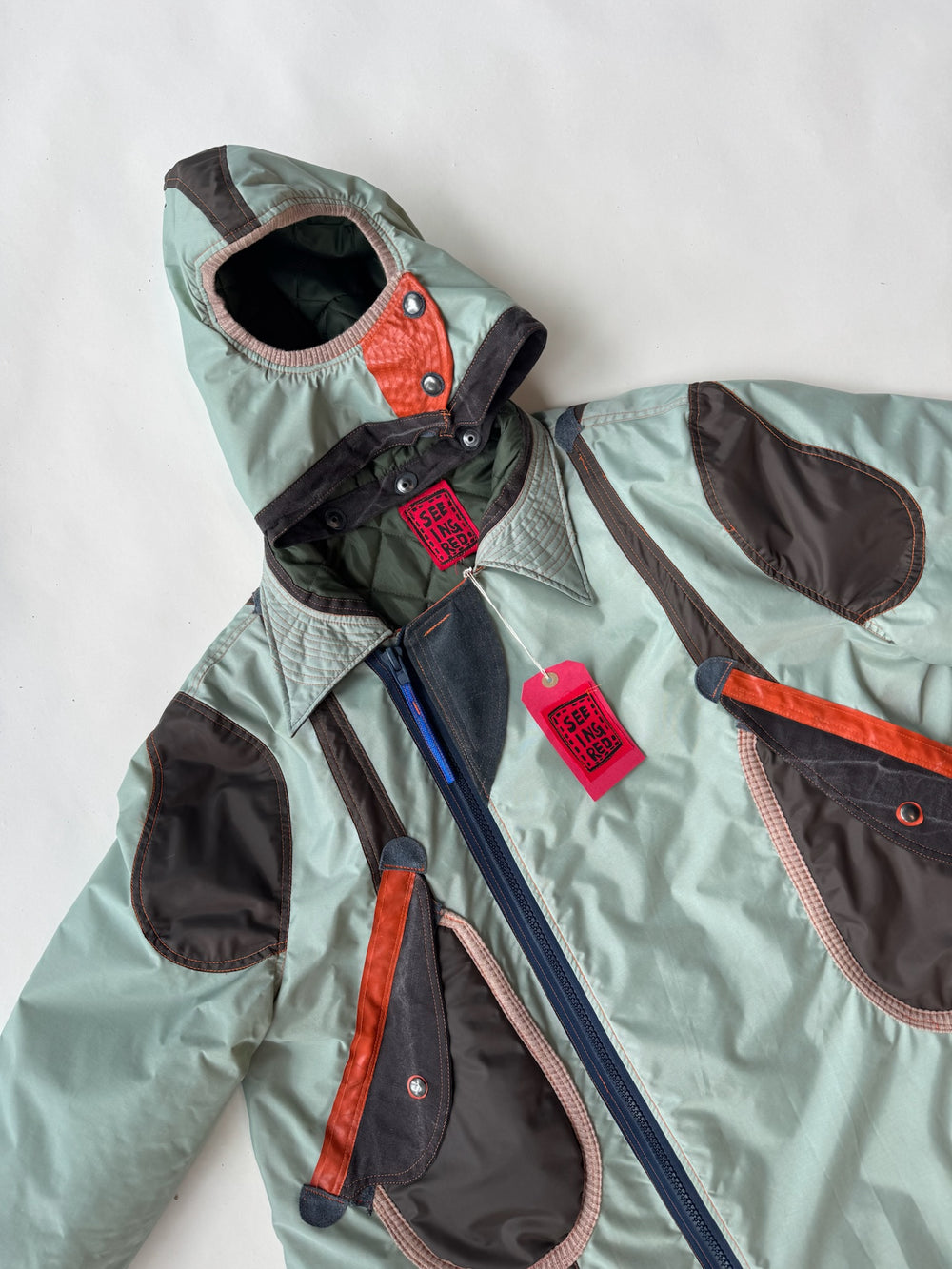 Mint green oversized parka with contrasting brown and red panels, featuring a sculptural hood with button details, asymmetrical pockets, and a bold red “I SEE” tag, laid flat on a white background.