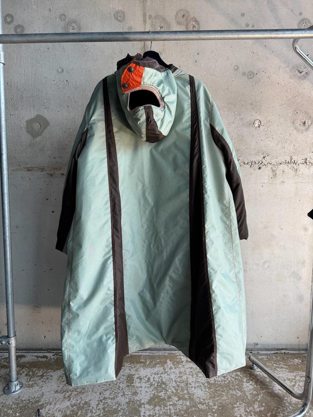 Back view of an oversized mint green parka with dark brown side panels and a structured hood featuring an orange quilted patch and black snap buttons; displayed on a hanger against a raw concrete backdrop.