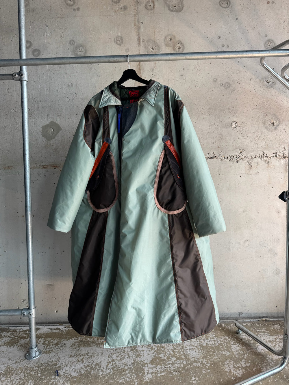 Unisex oversized mint green parka coat with dark brown geometric paneling, orange and grey accent details, and large sculptural flap pockets; displayed on a hanger against a concrete wall for an industrial aesthetic.