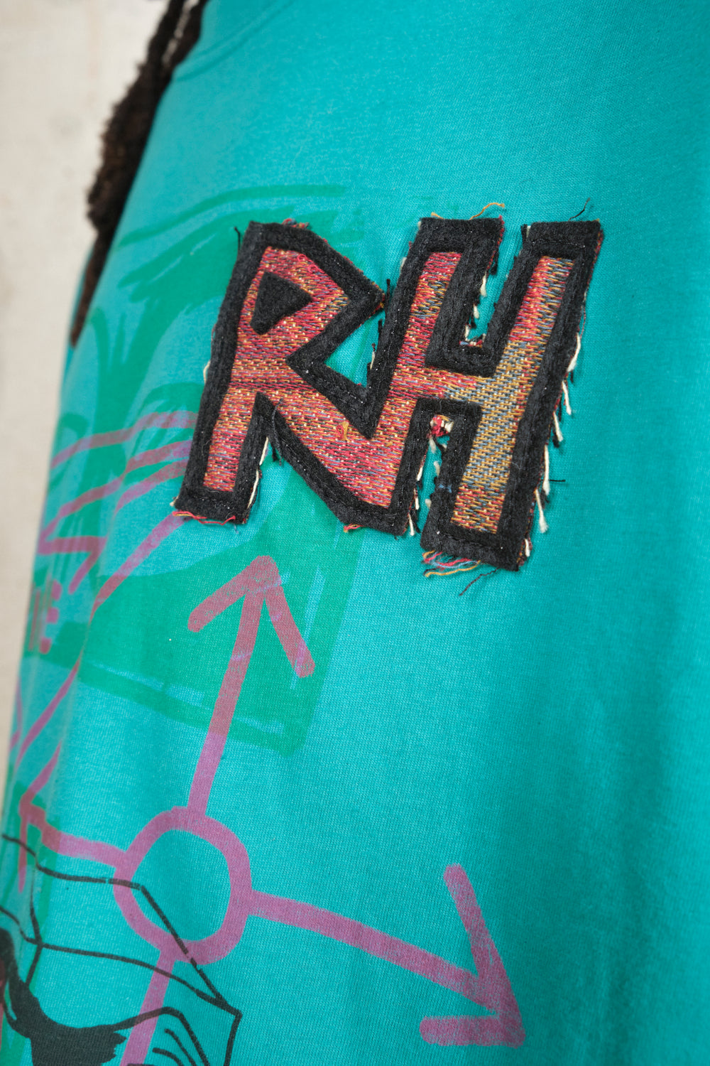 Close-up of textured RH patch in red and black thread on a turquoise T-shirt with colorful abstract graffiti design featuring arrows and eyes