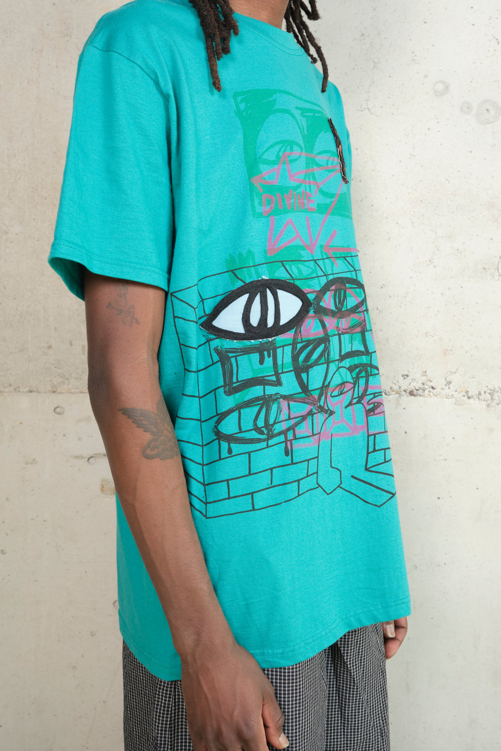 Side view of model wearing a turquoise graphic T-shirt with graffiti-inspired artwork of eyes, bricks, and the word ‘DIVINE’ in pink above a bold black-and-white embroidered eye.
