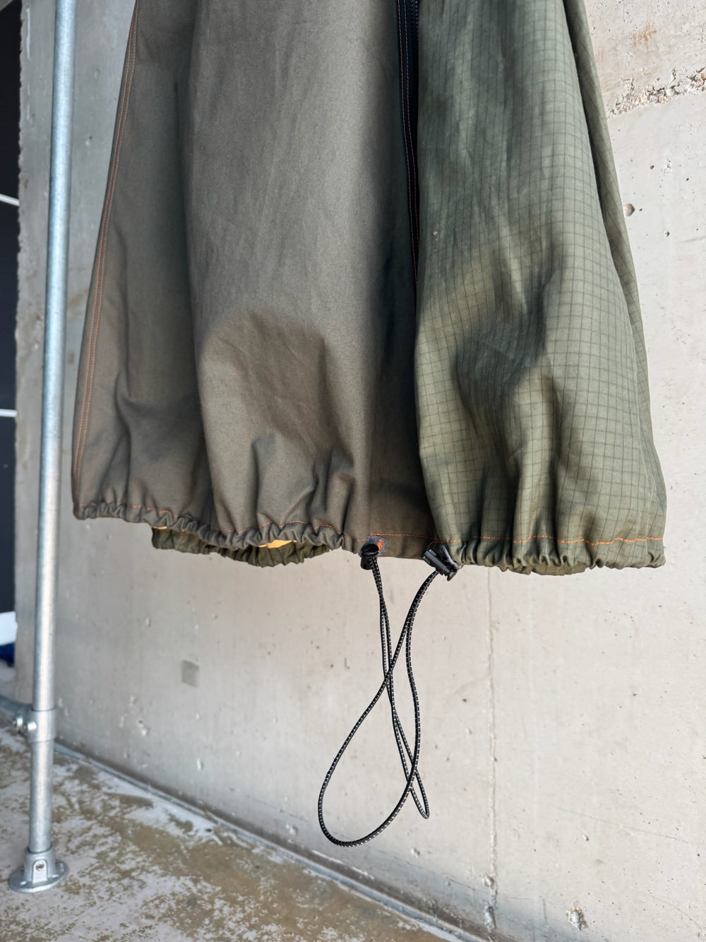 Close-up of the hem of a two-tone green skirt with ripstop panel, featuring an adjustable drawcord detail and gathered elastic bottom, photographed against a concrete background.