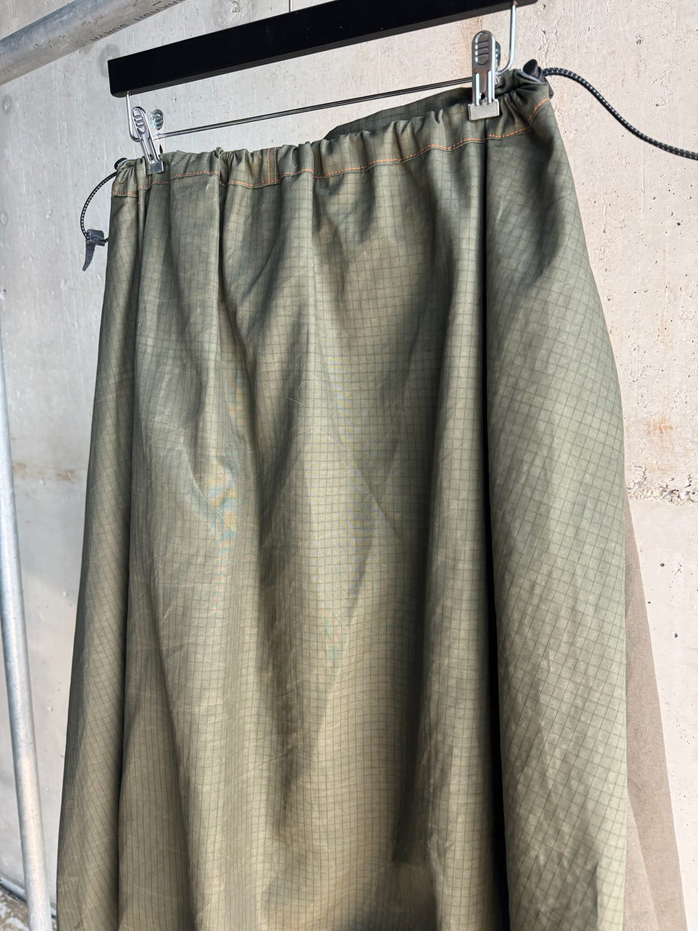 Close-up of the upper section of a green ripstop skirt with a gathered elastic waistband and visible orange topstitching, displayed on a hanger against a raw concrete