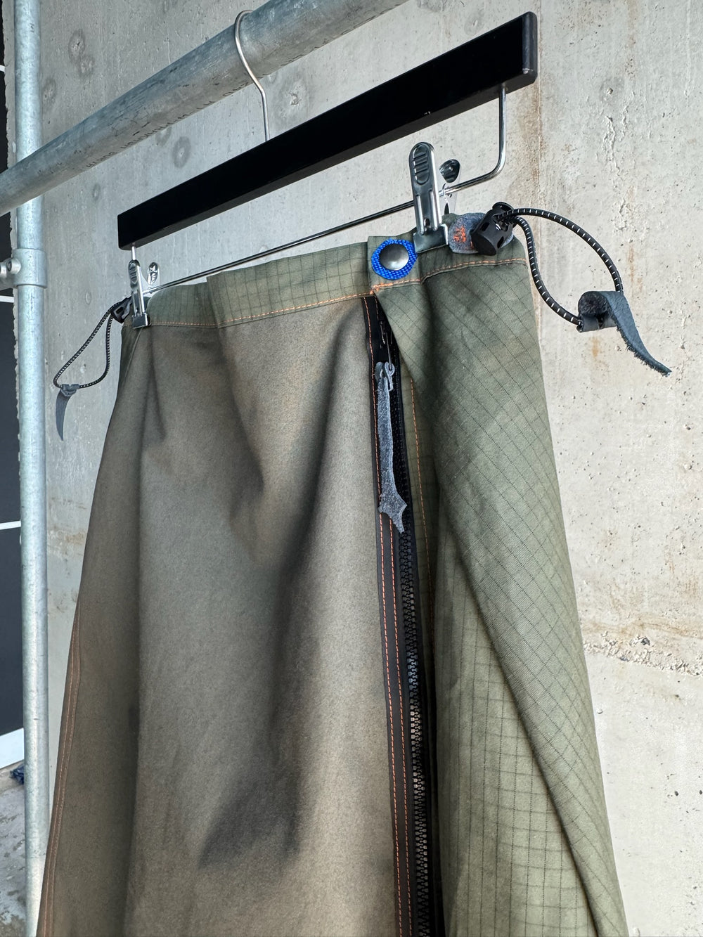 Close-up of a green asymmetrical skirt with ripstop panel detailing, black zipper with exposed teeth, and blue accent grommet on waistband, hanging from a black wooden hanger against a concrete wall.
