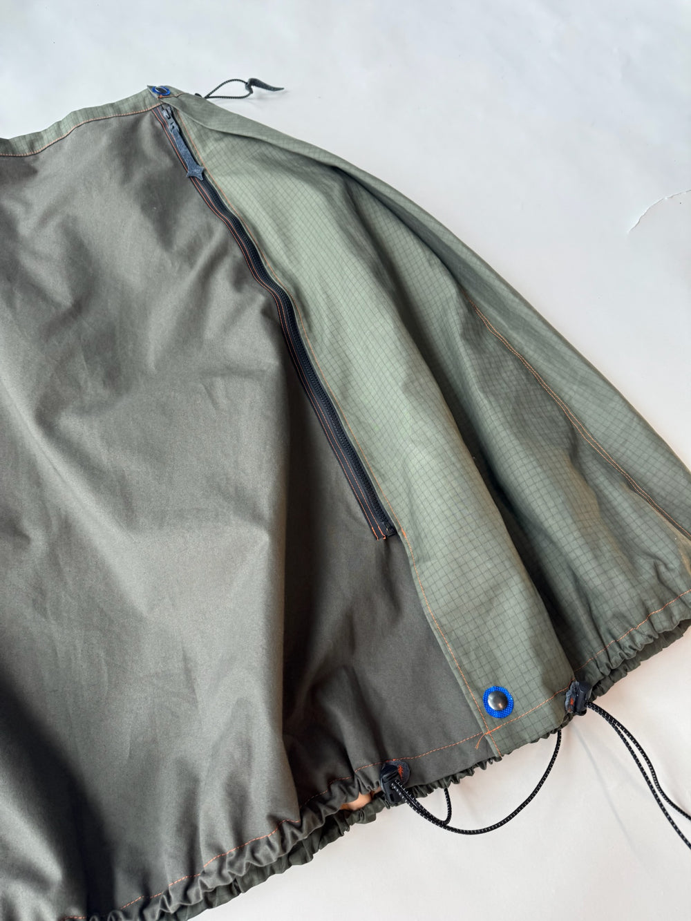 Close-up of an asymmetrical olive green skirt featuring contrasting ripstop fabric panels, a diagonal zipper, bright blue grommet detail, and an adjustable drawstring hem, laid flat on a light background.