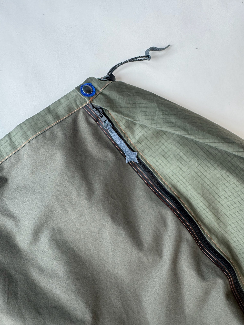 Close-up of olive green skirt waistband with contrasting ripstop panel, featuring a black zipper with custom pull tab, bright blue grommet, and adjustable drawcord detail on light background.