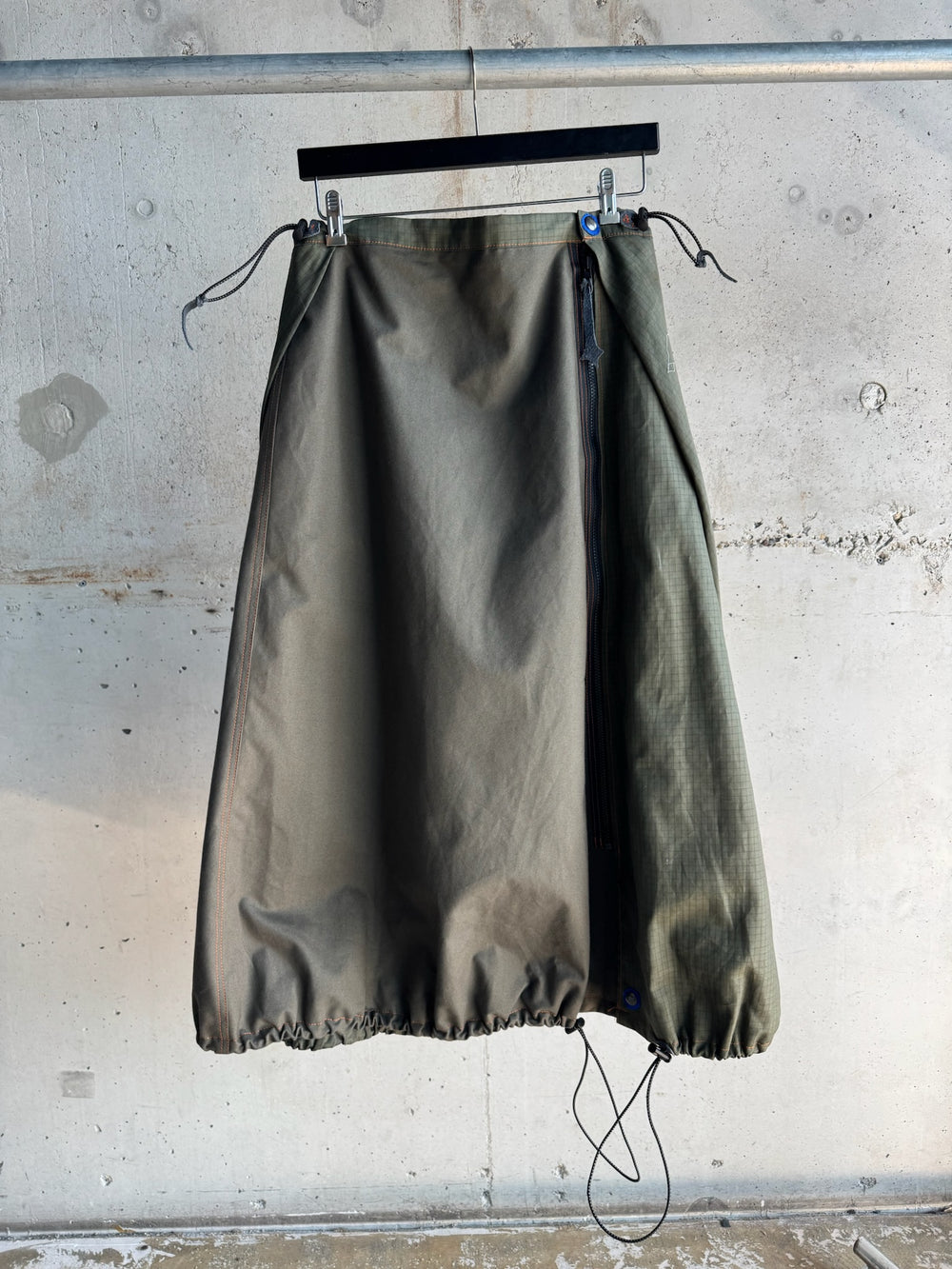 Army green A-line skirt with adjustable drawstrings at the hem, side zipper, and subtle orange topstitching, displayed on a hanger against a raw concrete wall backdrop.