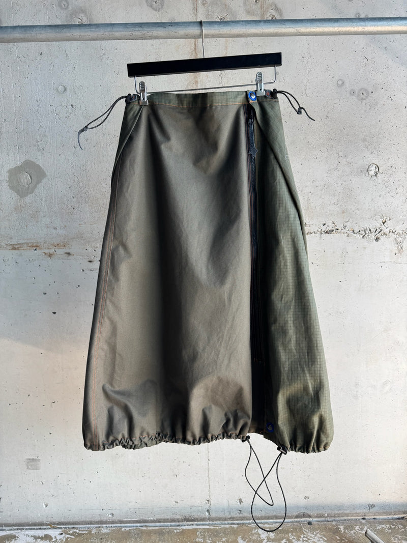 Army green A-line skirt with adjustable drawstrings at the hem, side zipper, and subtle orange topstitching, displayed on a hanger against a raw concrete wall backdrop.