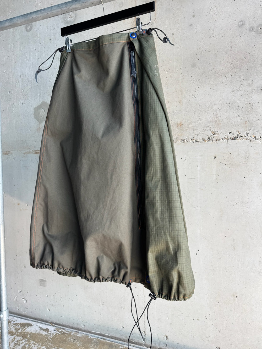 Asymmetric olive green skirt with zip closure and dual-texture panels, featuring ripstop fabric on the right side and drawstring hem, displayed hanging against a concrete wall backdrop.