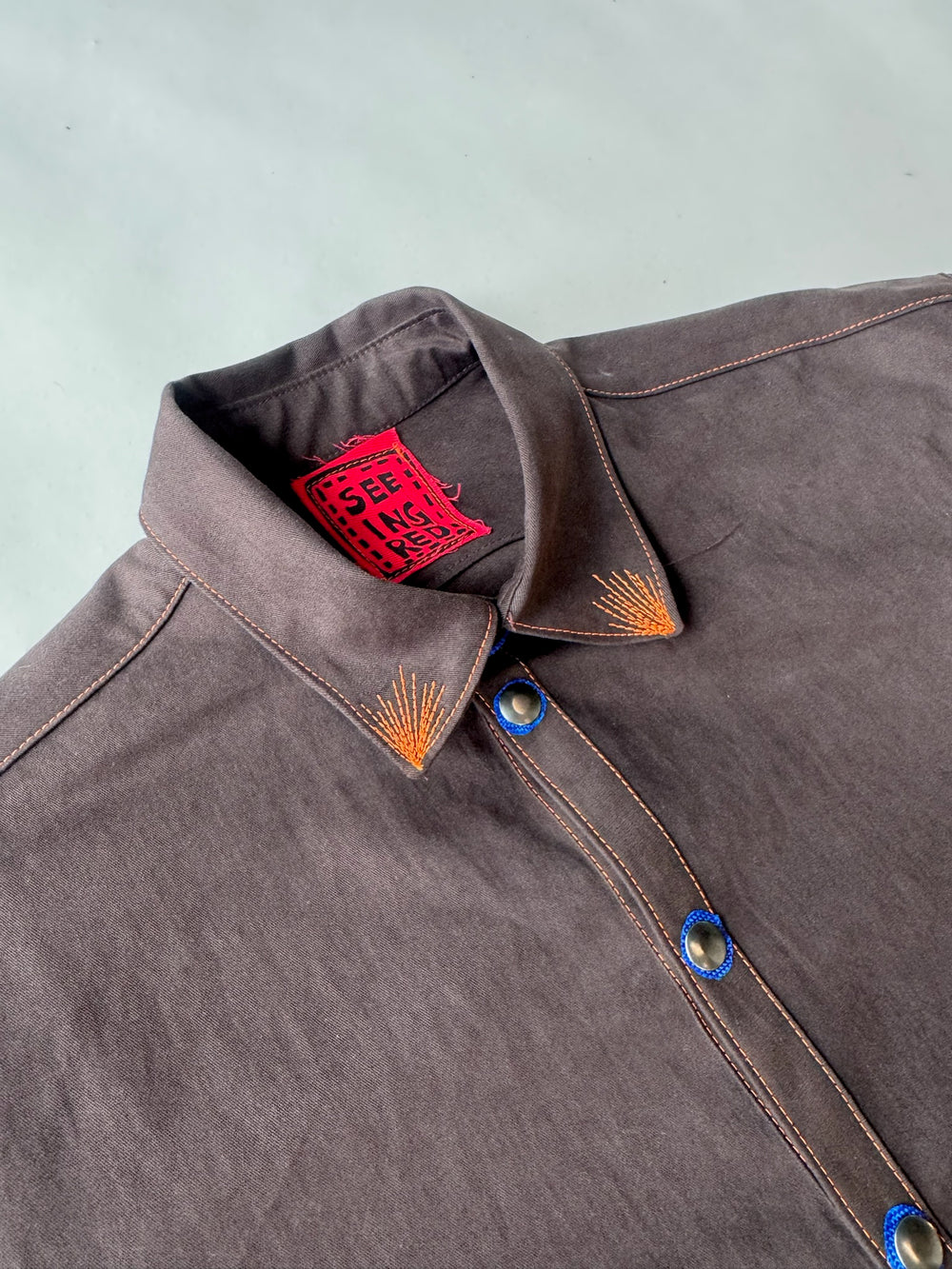 Close-up of a dark brown shirt featuring vibrant orange topstitching, blue-edged snap buttons, and a red inner label that reads “SEEING RED,” laid flat against a pale background.