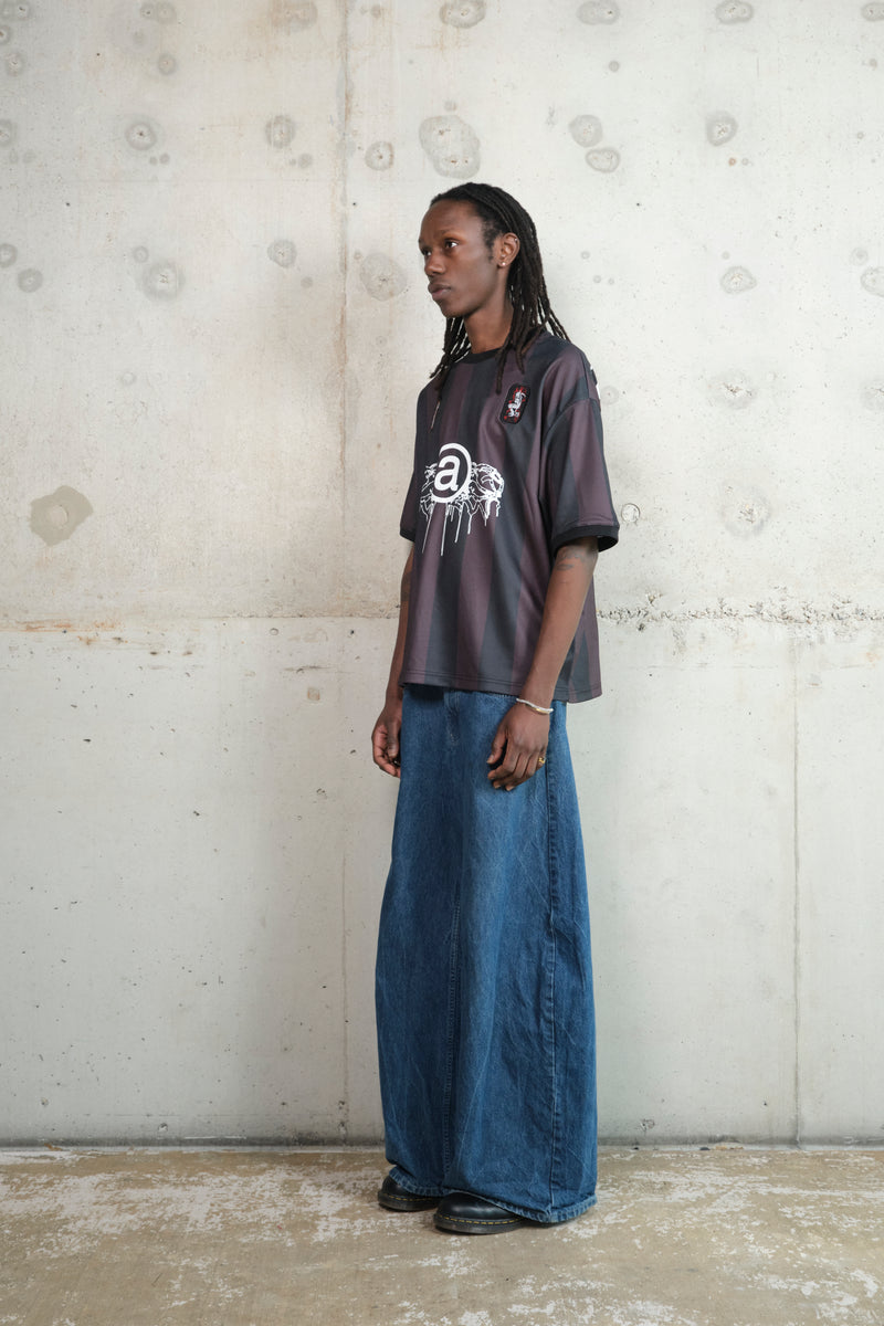 A person with long dreadlocks stands against a raw concrete wall, wearing an oversized black and maroon striped jersey with a bold white “@” symbol and dripping graphic design on the front. Small embroidered patches add detail to the shoulders. The jersey is paired with wide-leg blue denim jeans and black shoes, creating a casual yet statement look.