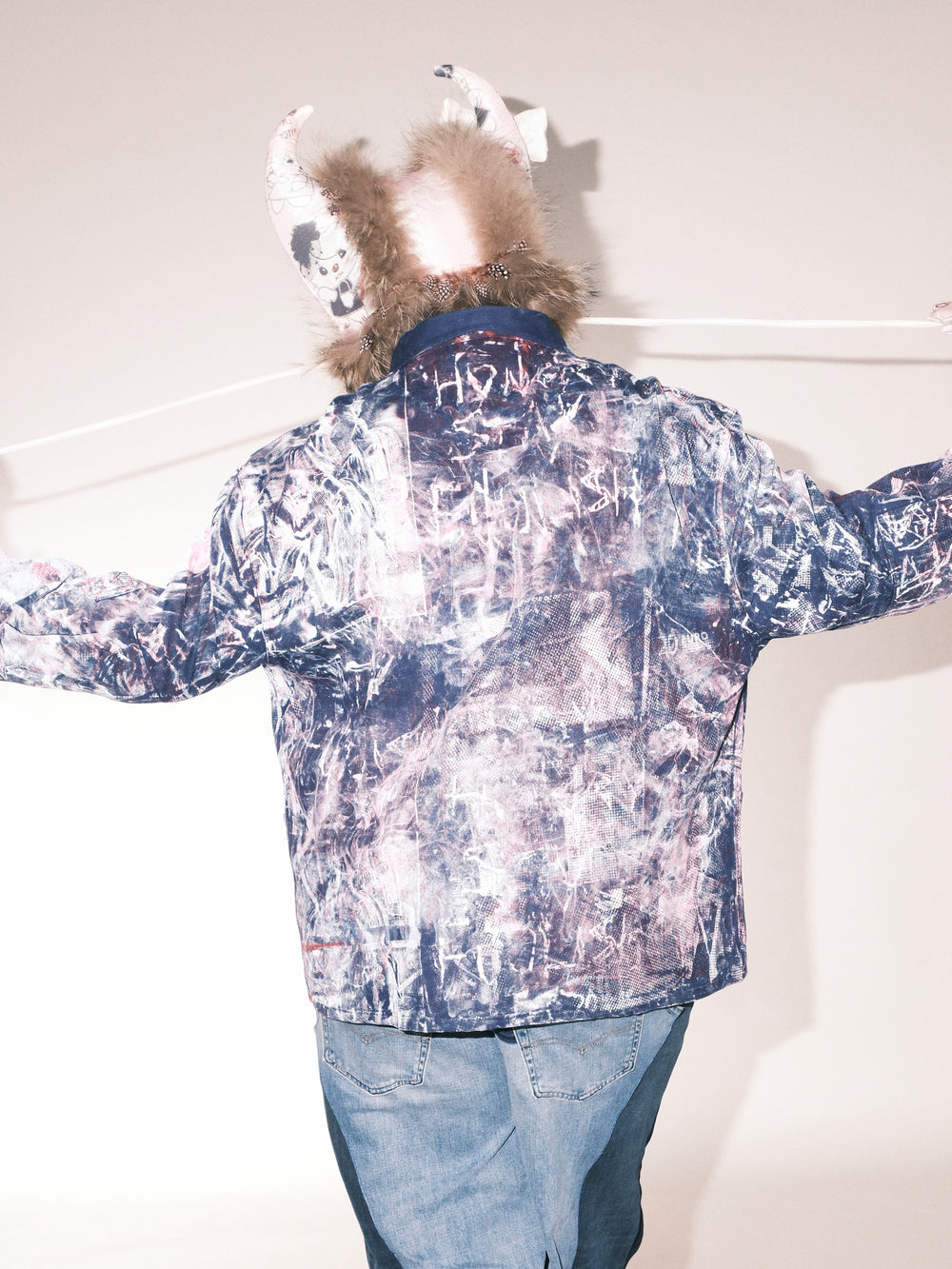 “Back view of a model wearing an abstract-patterned jacket in blue and pink tones with handwritten-style details. The outfit is paired with two-tone wide-leg denim pants and black boots. The model also wears a fur-lined hat with decorative ear-like elements.”