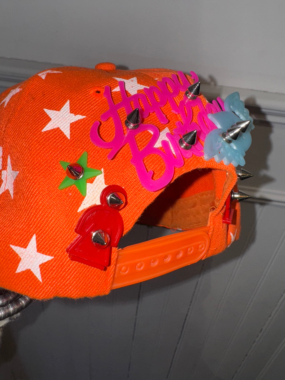 Close-up of the back of an orange baseball cap featuring a white star pattern, adorned with colorful embellishments, including a pink ‘Happy Birthday’ sign, spiked studs, a green star, and red and blue decorative elements. The adjustable snapback strap is visible at the bottom.