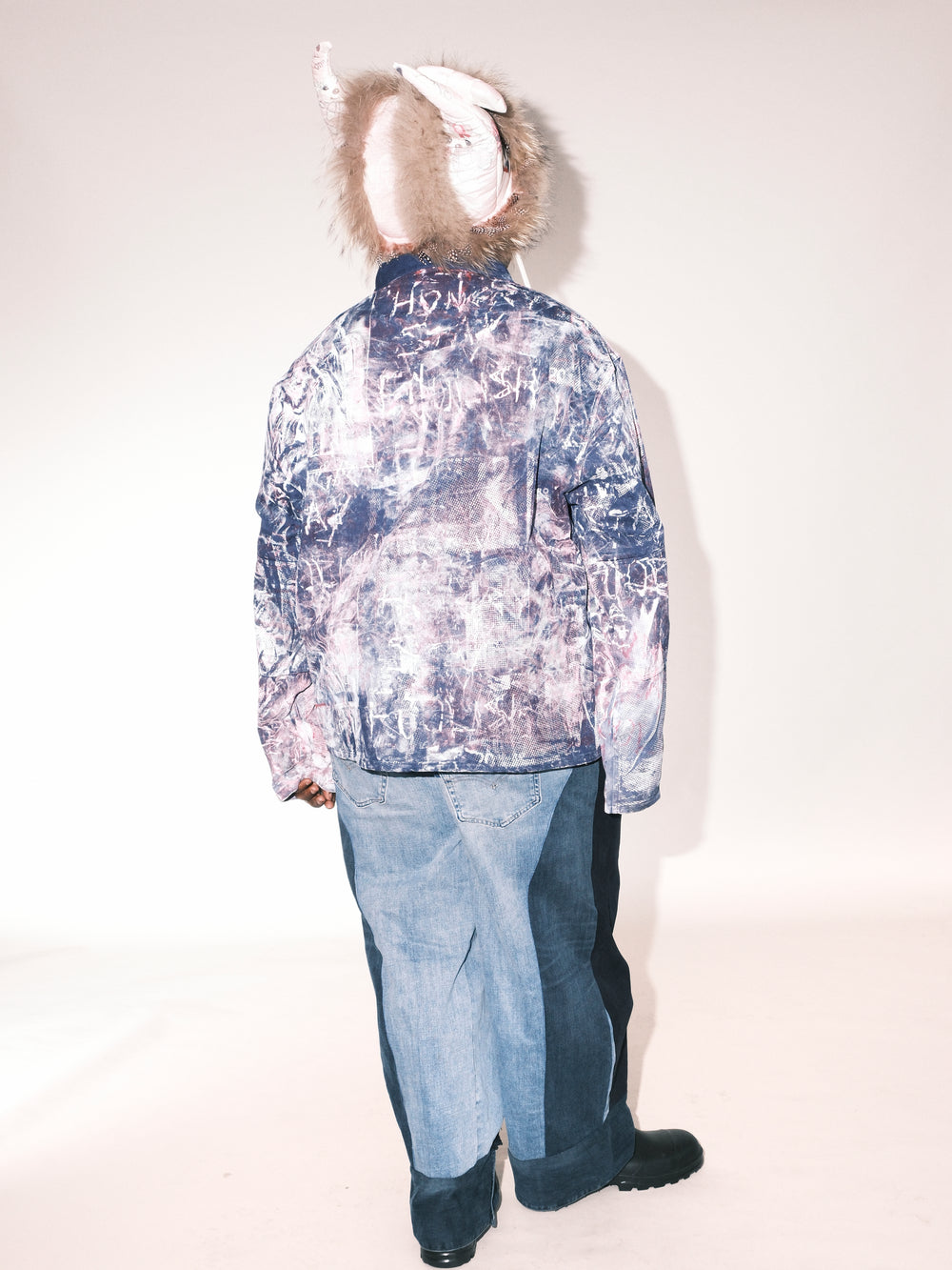 Back view of a model wearing an abstract-patterned jacket in blue and pink tones with handwritten-style details. The outfit is paired with two-tone wide-leg denim pants and black boots. The model also wears a fur-lined hat with decorative ear-like elements.