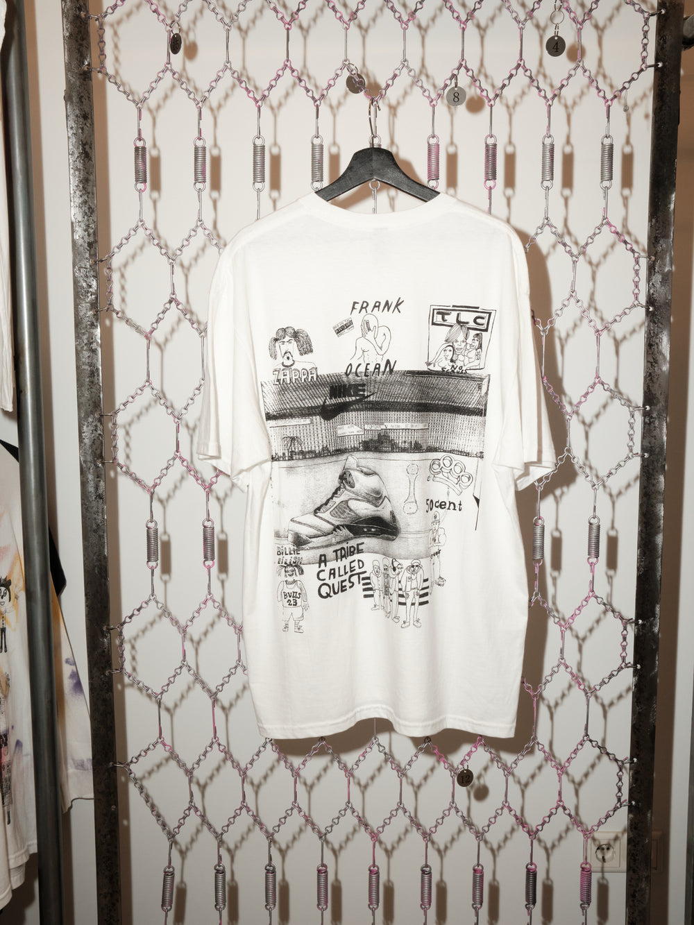 A white graphic T-shirt hangs on a black hanger against a chain-link patterned backdrop. The shirt features hand-drawn, collage-style black illustrations and text, including references to music artists such as Frank Ocean, TLC, Zappa, and A Tribe Called Quest, along with sketches of a sneaker and various cartoon-like figures