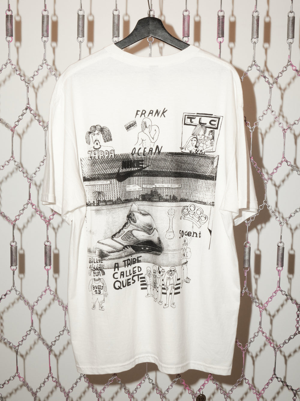 A white graphic T-shirt hangs on a black hanger against a chain-link patterned backdrop. The shirt features hand-drawn, collage-style black illustrations and text, including references to music artists such as Frank Ocean, TLC, Zappa, and A Tribe Called Quest, along with sketches of a sneaker and various cartoon-like figures