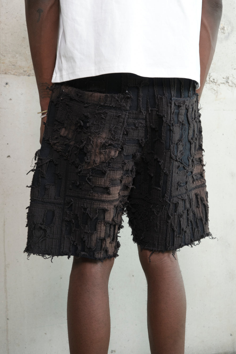 Close-up of a pair of black textured shorts with a deconstructed, patchwork design featuring frayed edges and layered fabric details. The model pairs them with a white oversized T-shirt, white crew socks, and black leather shoes. The background is a raw concrete wall.