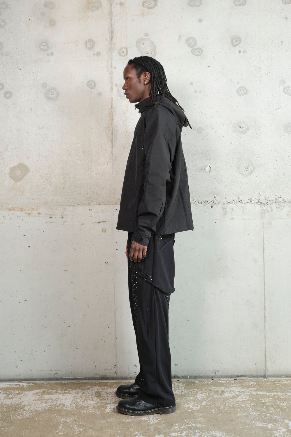 A male model with dark skin and long dreadlocks stands in profile against a textured concrete wall. He is wearing a black technical jacket with multiple zippered pockets and structured paneling, paired with black wide-leg pants featuring intricate silver stitching details. He completes the look with black leather shoes. His expression is neutral, and the minimalist, industrial background emphasizes the modern, streetwear-inspired aesthetic of the outfit.