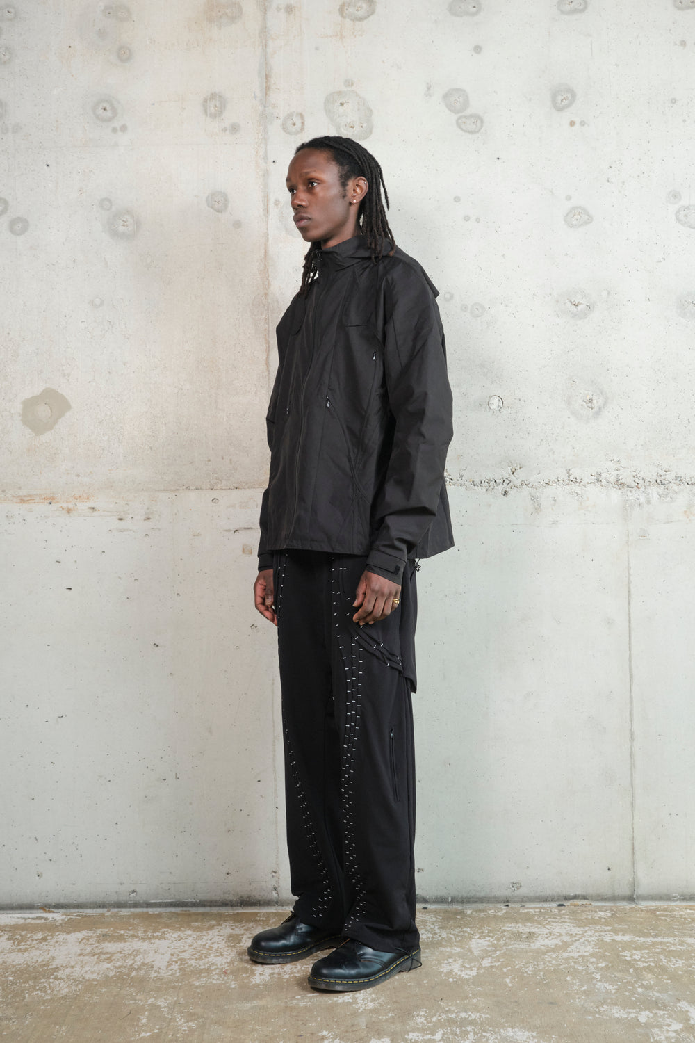A male model with dark skin and long dreadlocks stands in profile against a textured concrete wall. He is wearing a black technical jacket with multiple zippered pockets and structured paneling, paired with black wide-leg pants featuring intricate silver stitching details. He completes the look with black leather shoes. His expression is neutral, and the minimalist, industrial background emphasizes the modern, streetwear-inspired aesthetic of the outfit.