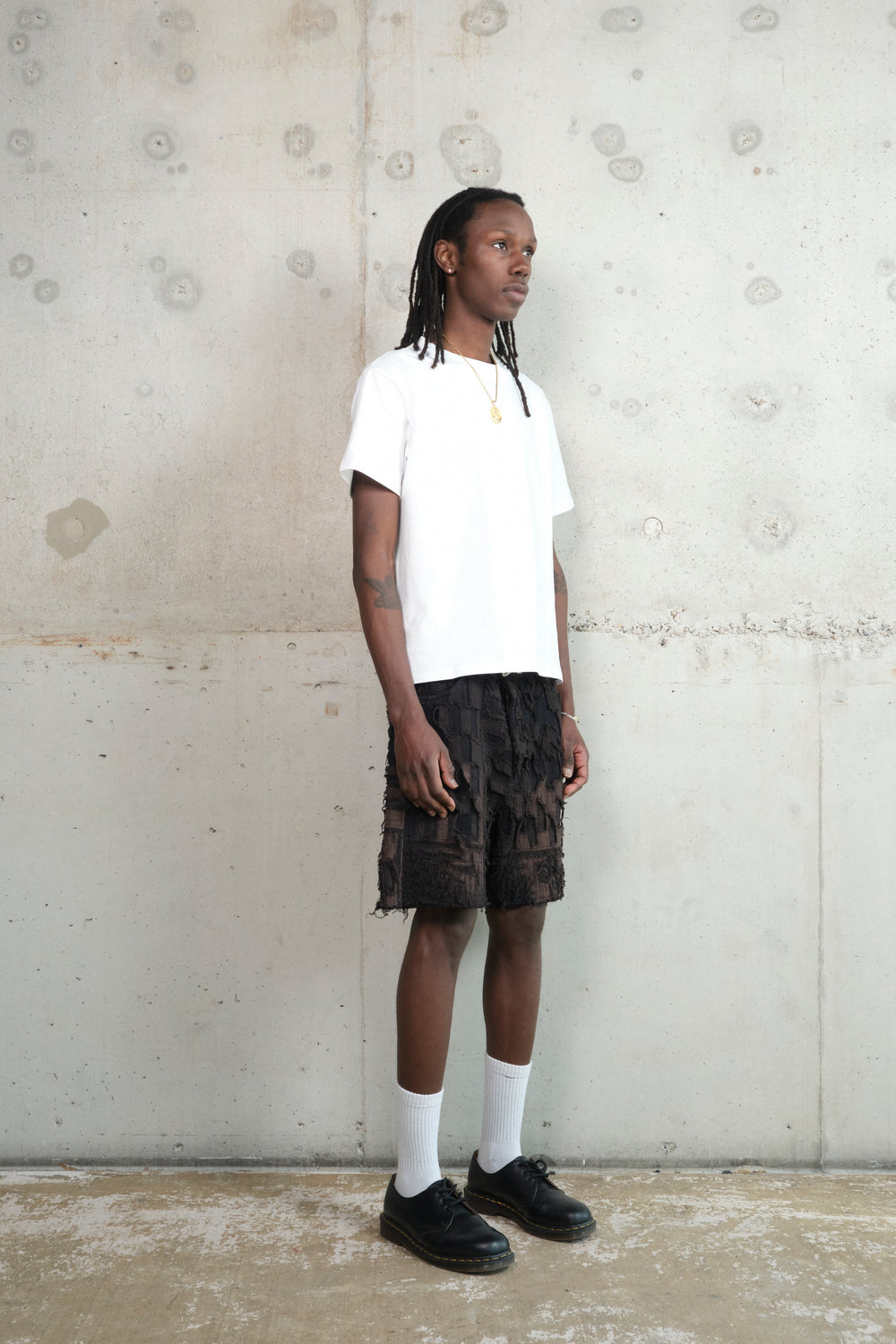 Model in a relaxed pose, wearing a minimalist outfit featuring a white oversized T-shirt, black textured shorts with a deconstructed design, white socks, and black leather shoes. Accessories include a gold pendant necklace and rings. The model stands against a raw concrete wall.