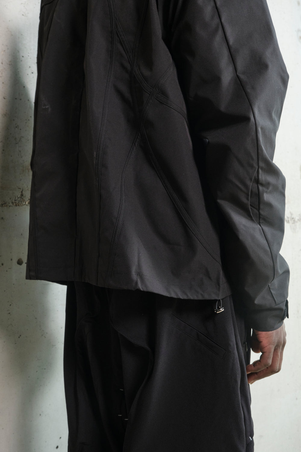 Close-up of a black technical jacket and pants, highlighting intricate seam detailing and structured paneling. The jacket features curved stitched lines and an adjustable drawcord at the hem. The pants include a discreet zippered pocket and layered fabric elements, adding depth to the design. The monochrome color scheme and tailored construction emphasize a modern, utilitarian aesthetic.