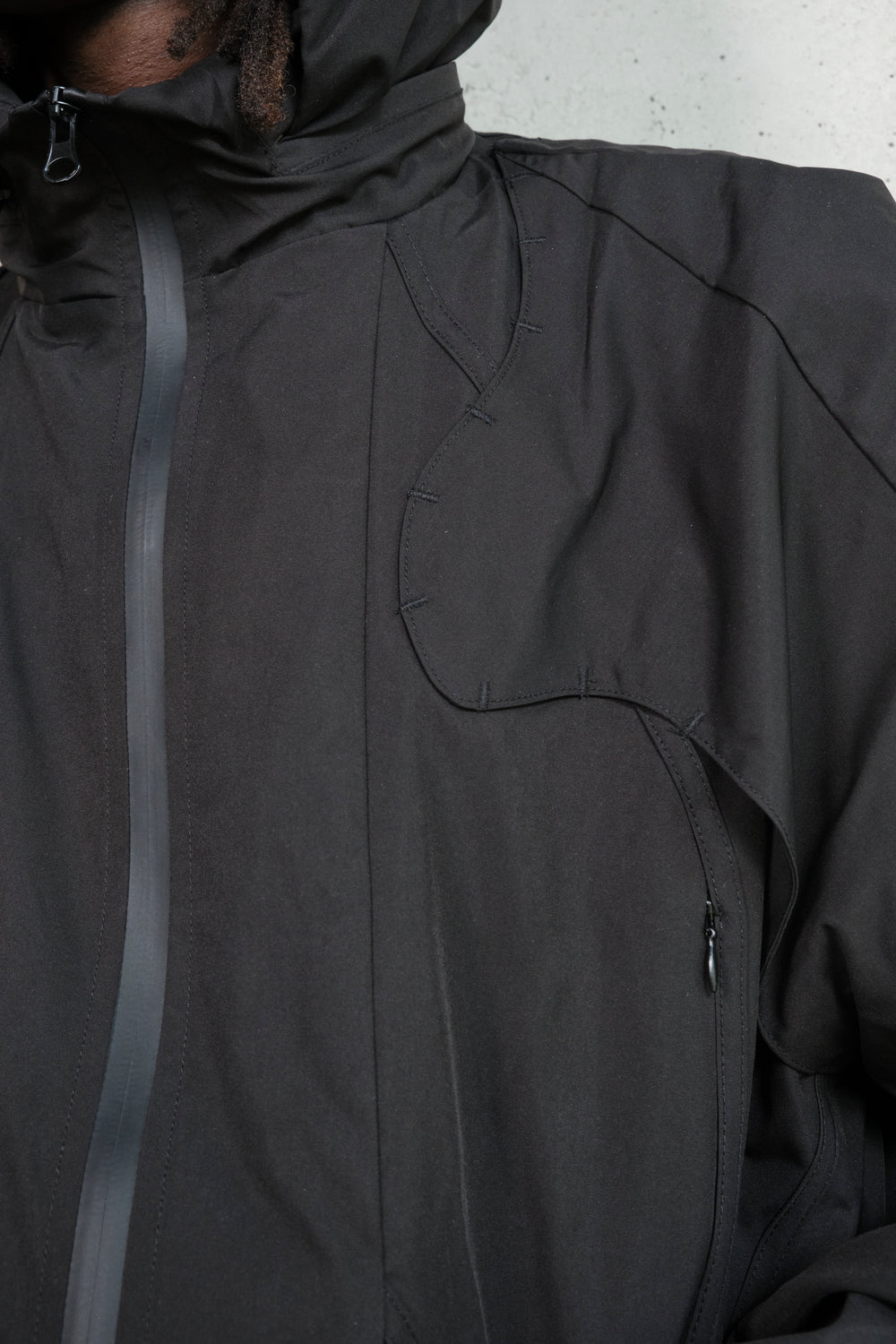 Close-up of a black technical jacket, highlighting intricate stitching details, a curved seam design on the chest, and a waterproof taped zipper. The fabric has a matte finish, and the precision of the construction emphasizes a modern, functional aesthetic. A small zippered pocket is visible on the lower part of the jacket, enhancing its utility.