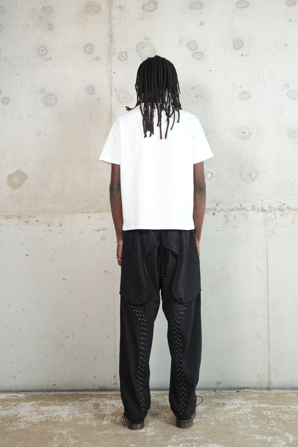 A male model with long dreadlocks stands with his back facing the camera against a minimalist concrete wall. He wears a white oversized T-shirt and black wide-leg pants with silver stitching details. The back of the pants features layered fabric panels, adding depth and movement to the design. His stance is relaxed, highlighting the effortless, modern aesthetic of the outfit.