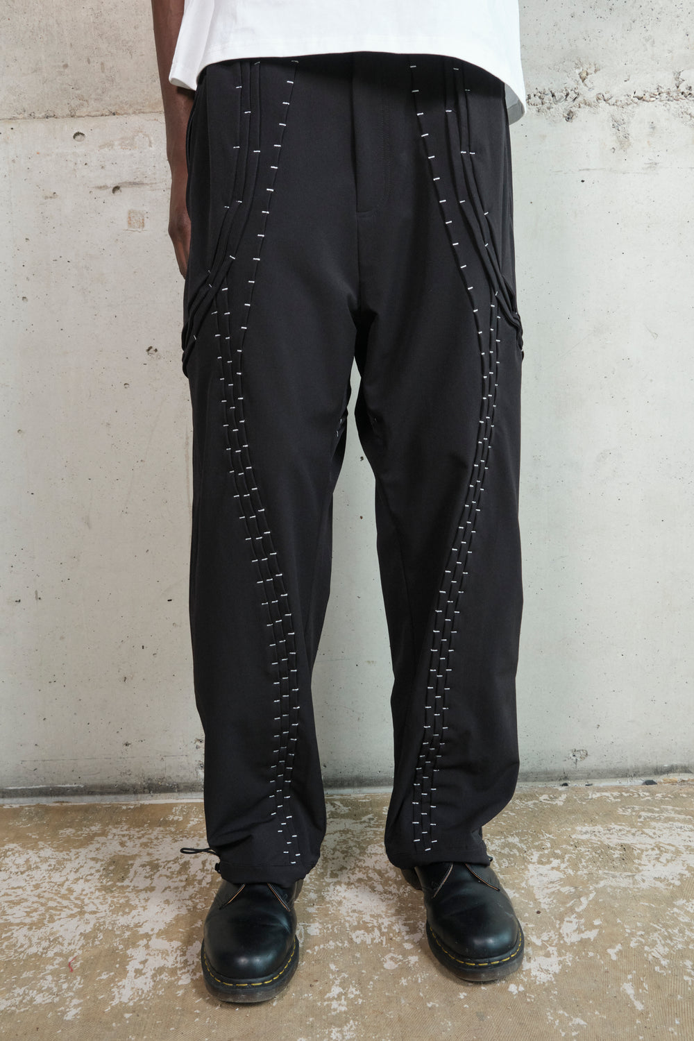 Close-up of black wide-leg pants featuring intricate silver stitching details running vertically along the legs. The pants have structured paneling and zippered pockets on the sides, adding a modern and utilitarian touch. The model pairs them with black leather shoes, enhancing the edgy, streetwear-inspired aesthetic.