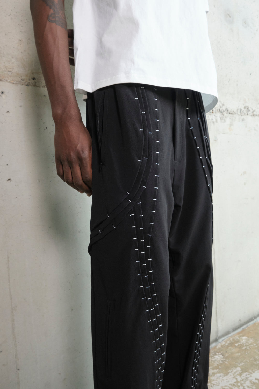 Close-up of black wide-leg pants with curved silver stitching details and layered paneling. The pants feature zippered pockets and structured seams, adding depth and dimension to the design. The model is wearing a crisp white T-shirt, creating a strong contrast against the dark tones of the pants. The composition highlights the craftsmanship and modern aesthetic of the garment.7uhgfr