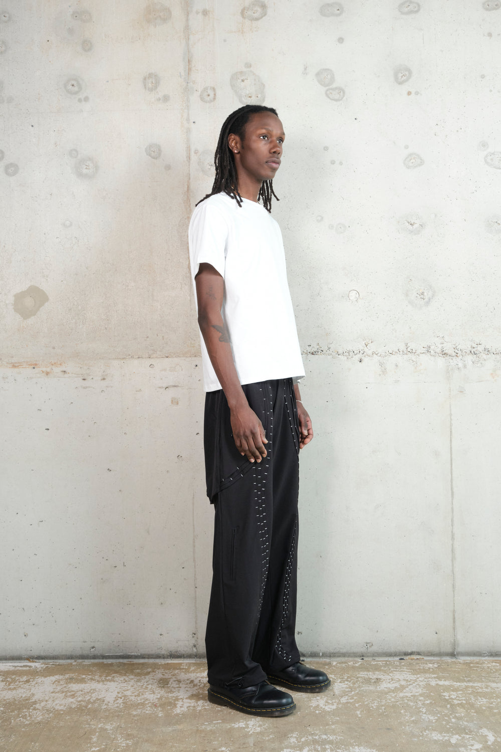 A male model with dark skin and long dreadlocks stands in a relaxed pose against a textured concrete wall. He wears a white oversized T-shirt paired with black wide-leg pants featuring silver stitching details. His black leather shoes complete the monochrome ensemble. The side profile highlights the clean lines and minimalistic aesthetic of the outfit.