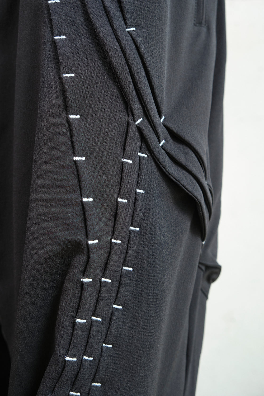 Close-up of black pants showcasing intricate white stitching details running vertically along layered fabric panels. The precise contrast stitching highlights the craftsmanship and structured design, adding depth and texture to the garment. The soft draping enhances the modern and avant-garde aesthetic.