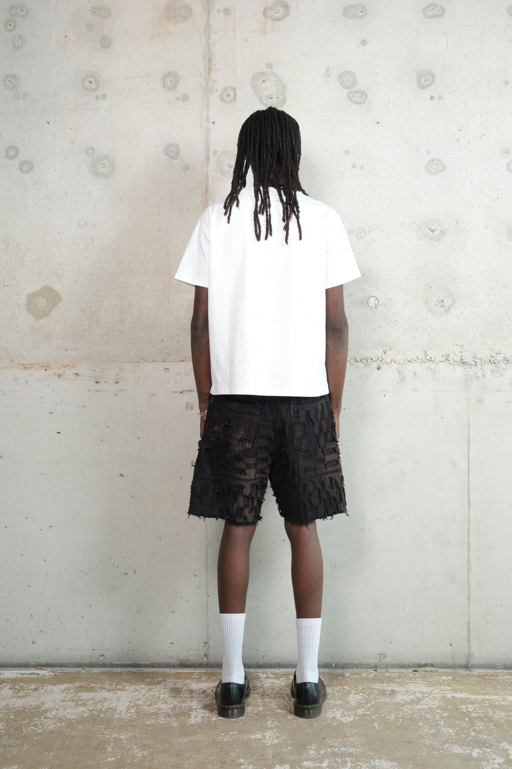 Model in a relaxed pose, wearing a minimalist outfit featuring a white oversized T-shirt, black textured shorts with a deconstructed design, white socks, and black leather shoes. Accessories include a gold pendant necklace and rings. The model stands against a raw concrete wall.