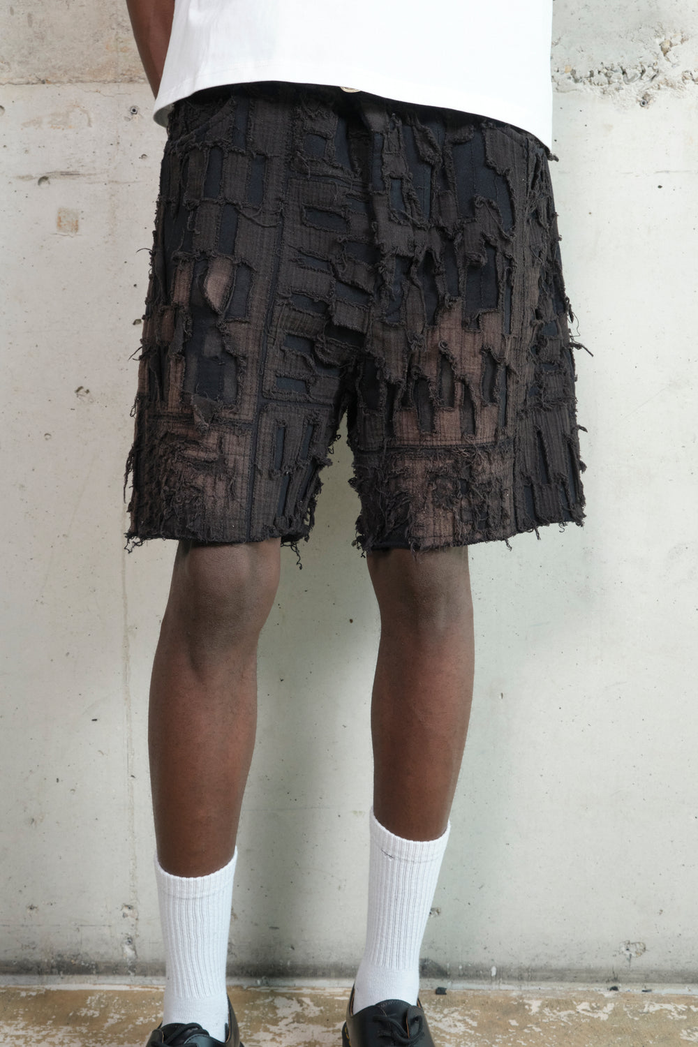 Close-up of a pair of black textured shorts with a deconstructed, patchwork design featuring frayed edges and layered fabric details. The model pairs them with a white oversized T-shirt, white crew socks, and black leather shoes. The background is a raw concrete wall.