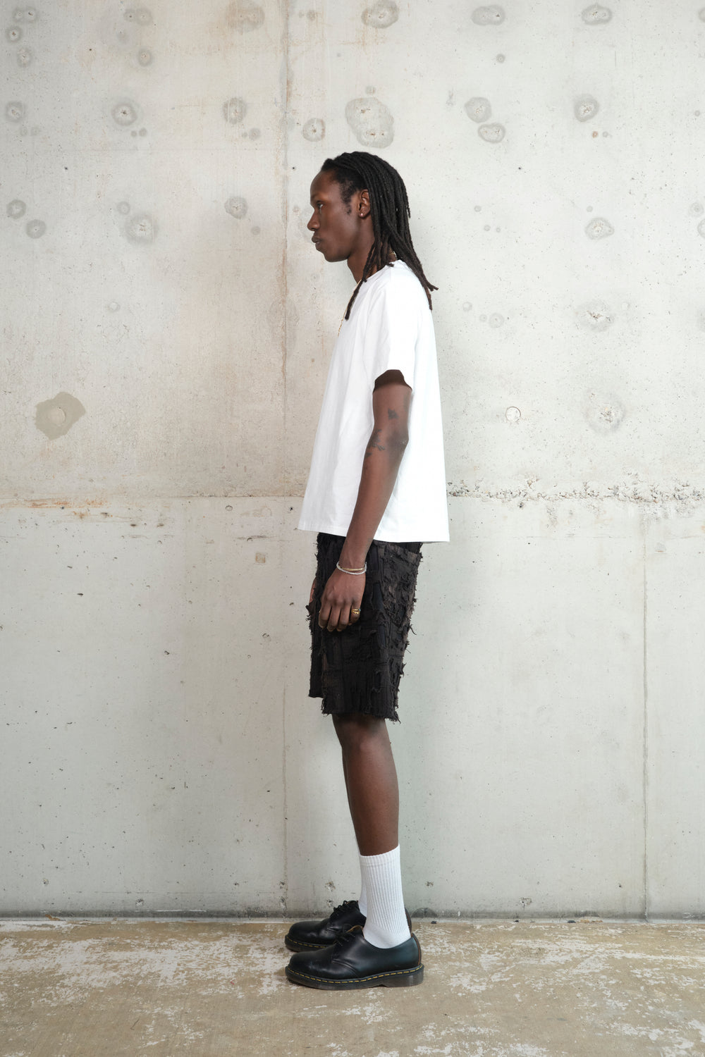 Model in a relaxed pose, wearing a minimalist outfit featuring a white oversized T-shirt, black textured shorts with a deconstructed design, white socks, and black leather shoes. Accessories include a gold pendant necklace and rings. The model stands against a raw concrete wall.