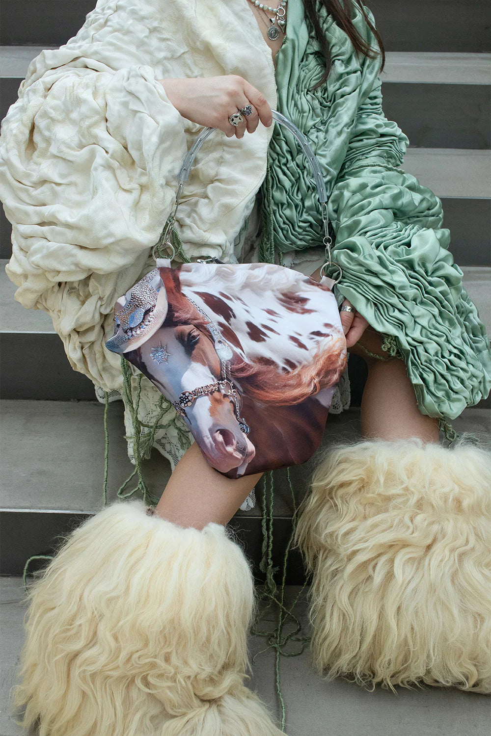 Person wearing a fluffy white coat and green scarf, holding a horse-print handbag.