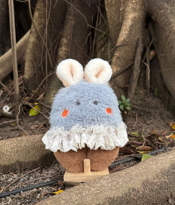 Handmade blue and brown bunny plush with lace skirt and floppy ears, posed on wooden stand among tree roots in natural forest setting – Mimi Planet collectible toy