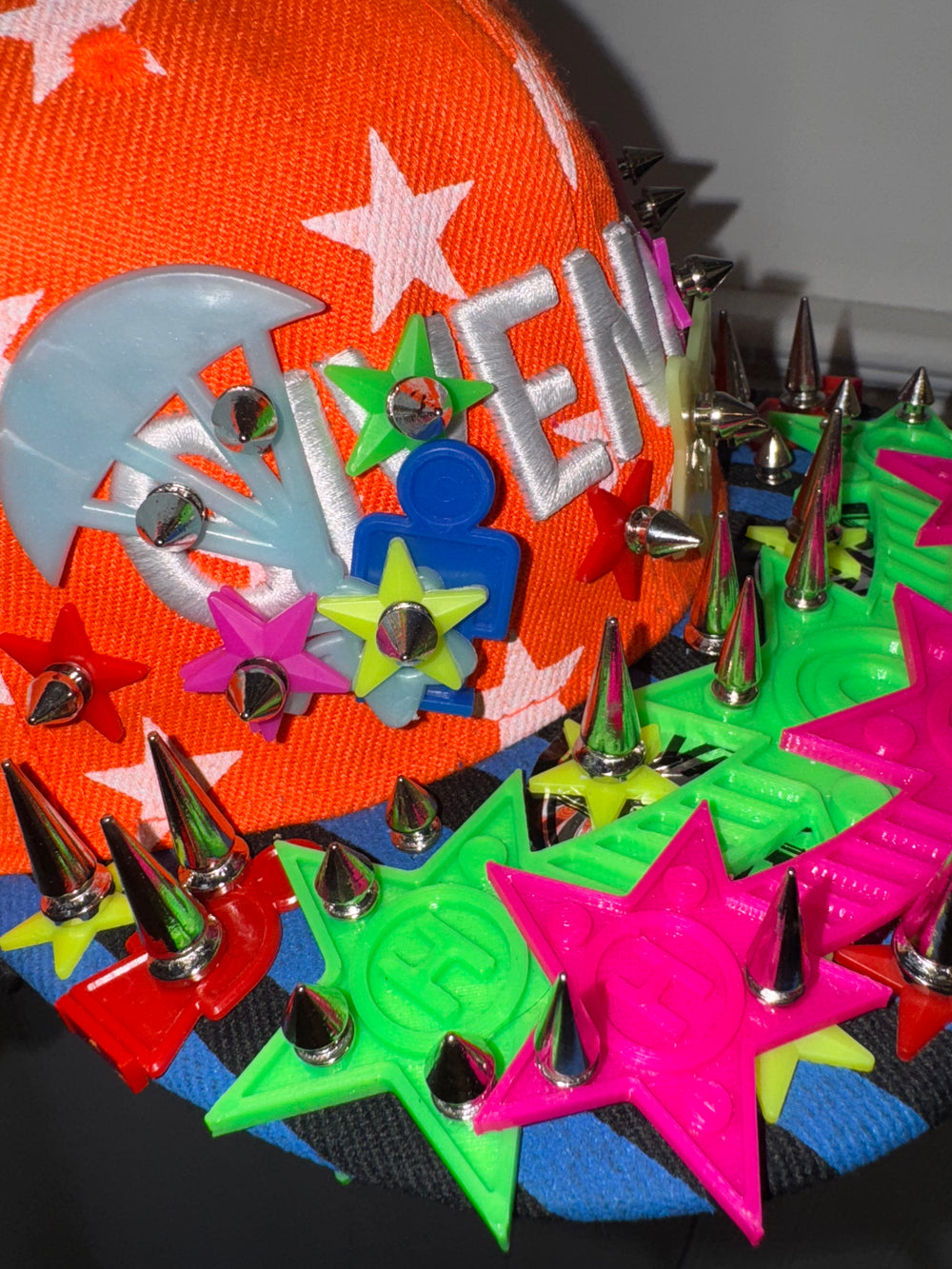 An orange baseball cap decorated with white stars and the word ‘VENCHY’ embroidered in white letters. The brim and front are adorned with colorful 3D embellishments, including bright pink and green star shapes, multicolored flower designs, and metallic silver spikes, creating a bold and playful aesthetic.