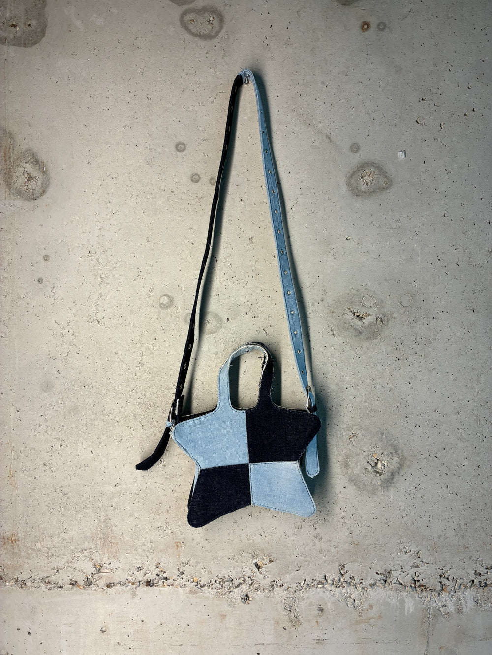 Back view of two-tone denim star-shaped shoulder bag with adjustable strap displayed on concrete wall.