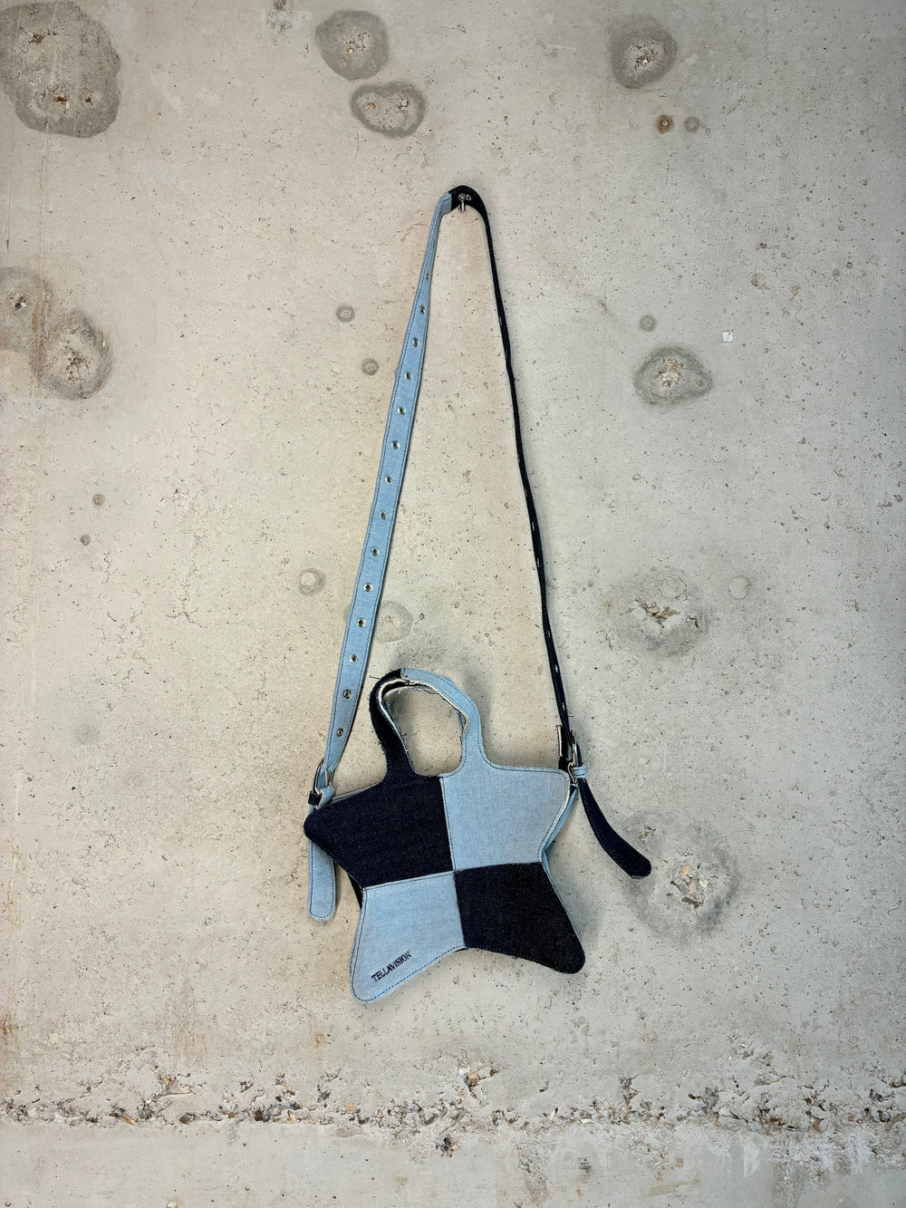 Patchwork denim star-shaped shoulder bag in light and dark blue wash with adjustable strap hanging against concrete wall.