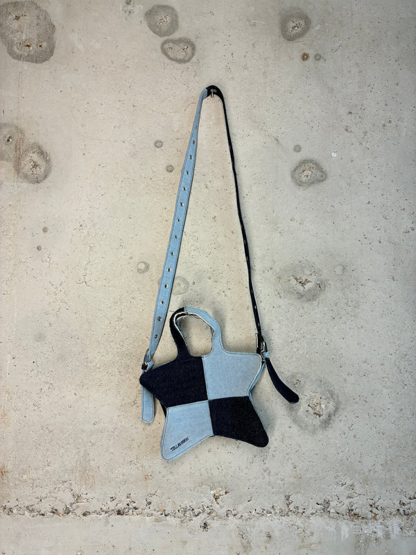 Patchwork denim star-shaped shoulder bag in light and dark blue wash with adjustable strap hanging against concrete wall.