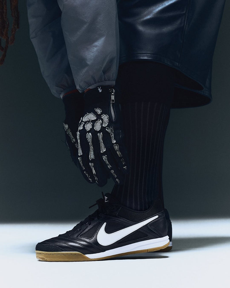 The image shows a close-up of a person wearing black Nike indoor soccer shoes with a white swoosh and gum soles. They are also wearing black skeleton-themed gloves, ribbed black socks, and an outfit featuring leather and synthetic materials. The setting appears to be a studio with soft lighting.
